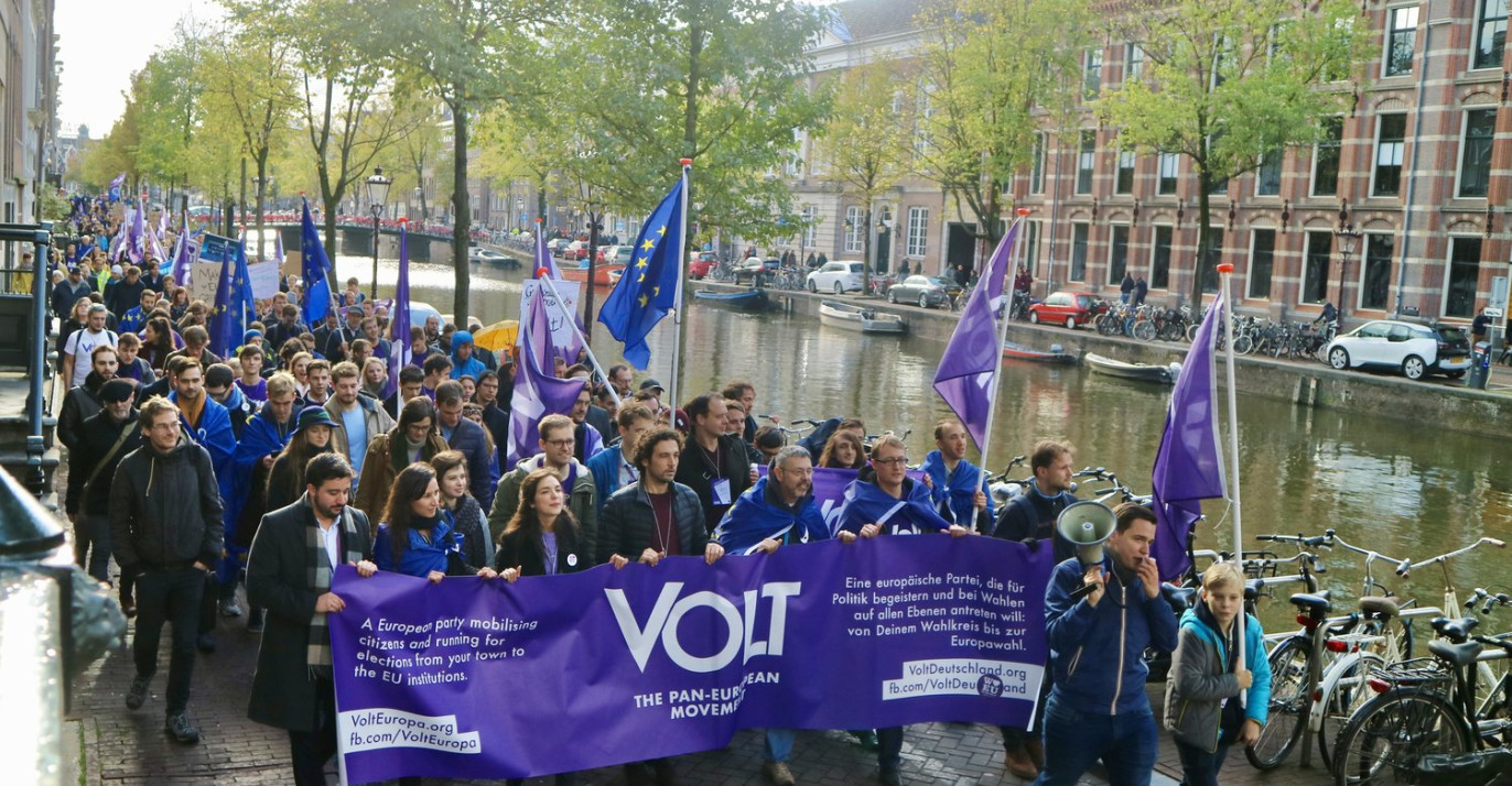 Een optocht van een grote groep Vlotters die lopen over straat. De hebben een grote banner voorop en houden vlaggen in de lucht.